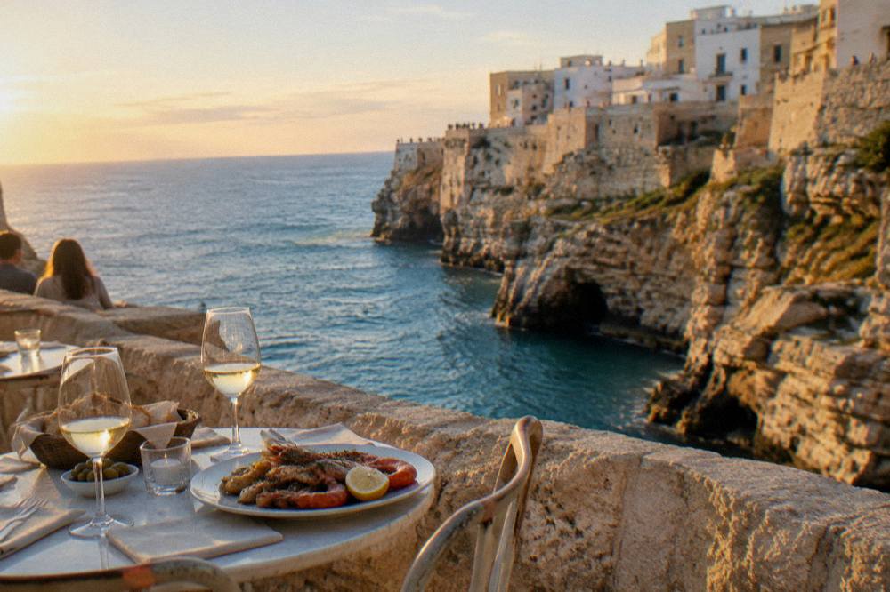 Small round tables on a cliff-edge terrace with glasses and a plate of seafood above blue water