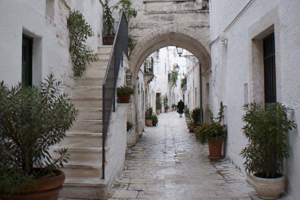 Narrow white old town lane with a stone arch and potted plants in afternoon shade