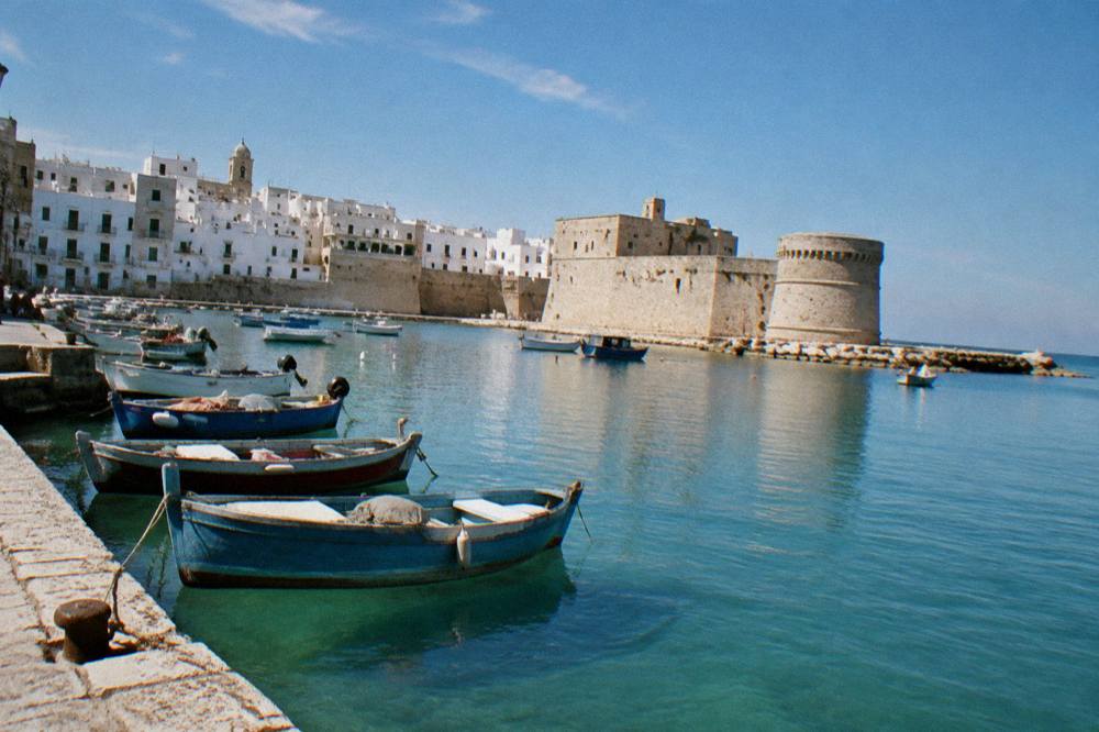 Old harbour of small painted wooden boats with a stone castle on the headland and a whitewashed old town behind