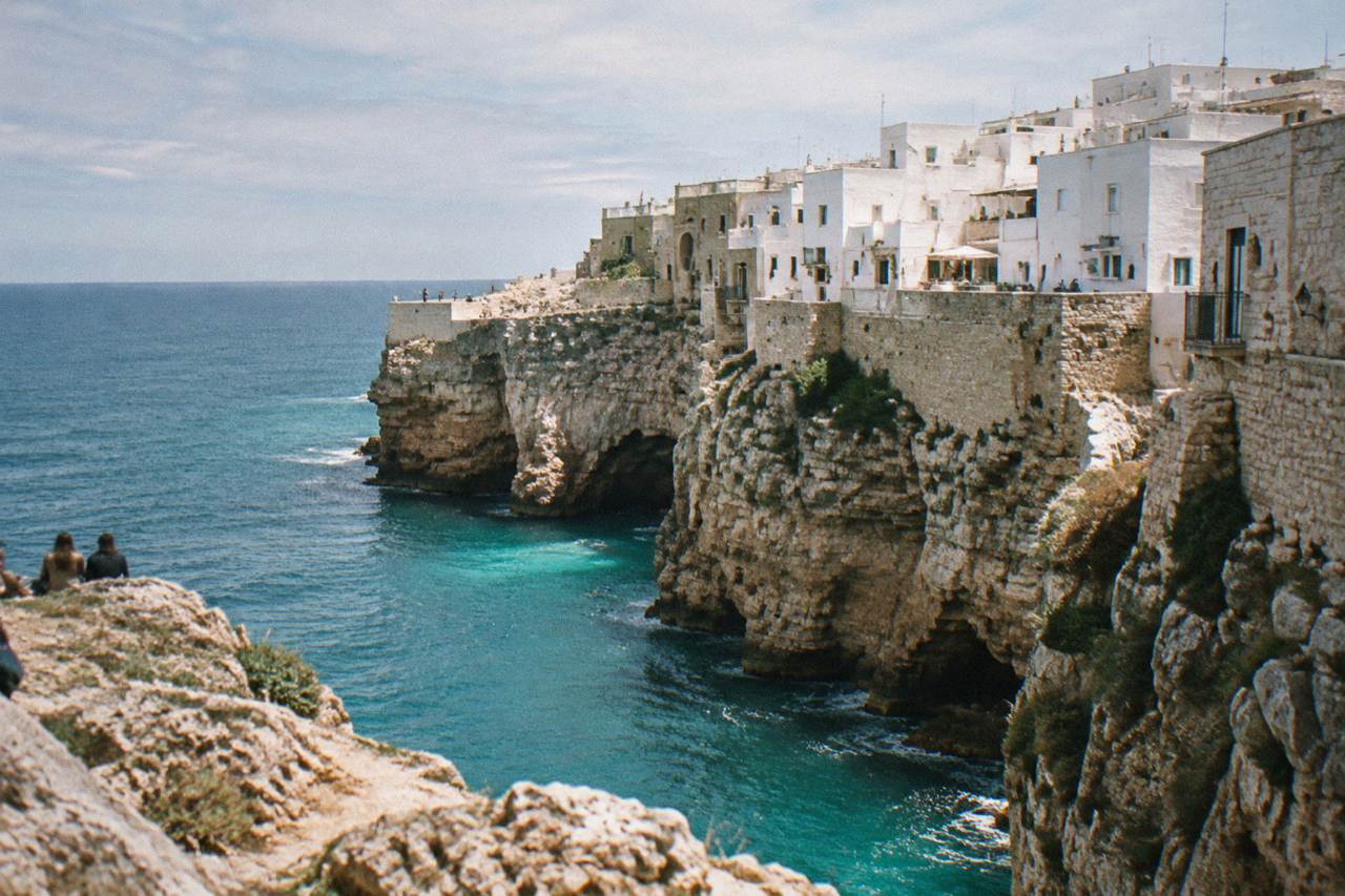White town perched on the edge of tall pale limestone cliffs above a turquoise sea seen from a rocky viewpoint