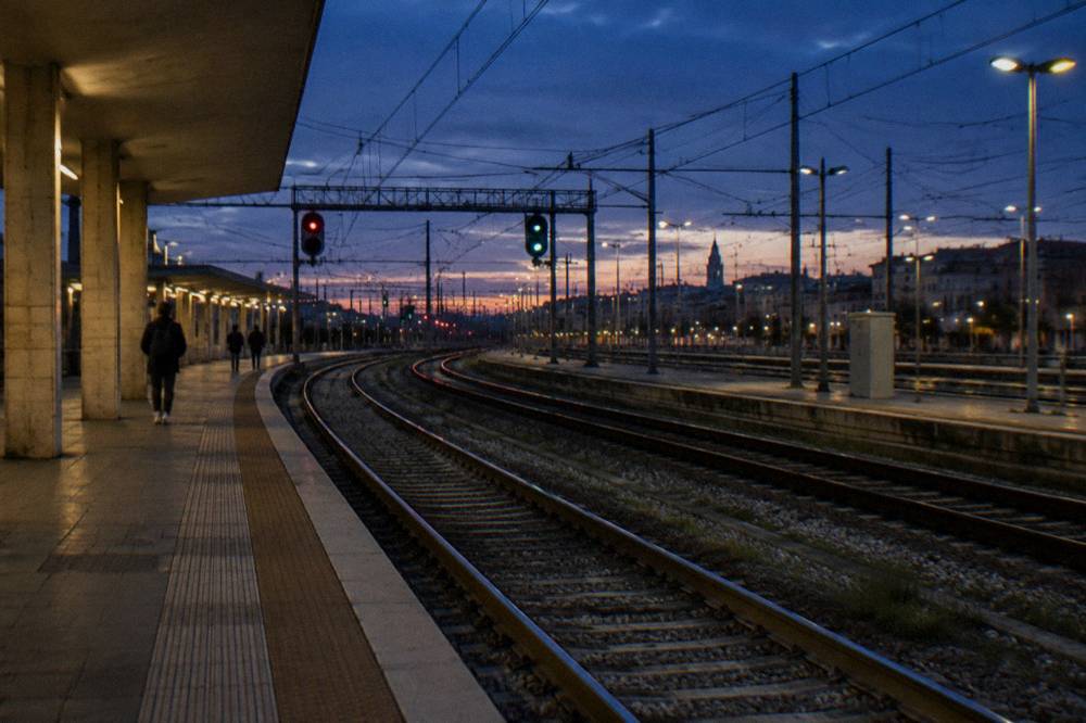 Rails curving away from a platform end at dusk with glowing signal lights and a few distant passengers
