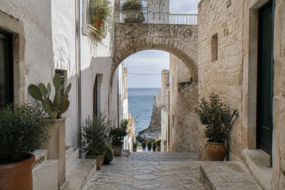 Narrow whitewashed cliff-top old town lane with a stone arch, potted plants and a glimpse of blue sea