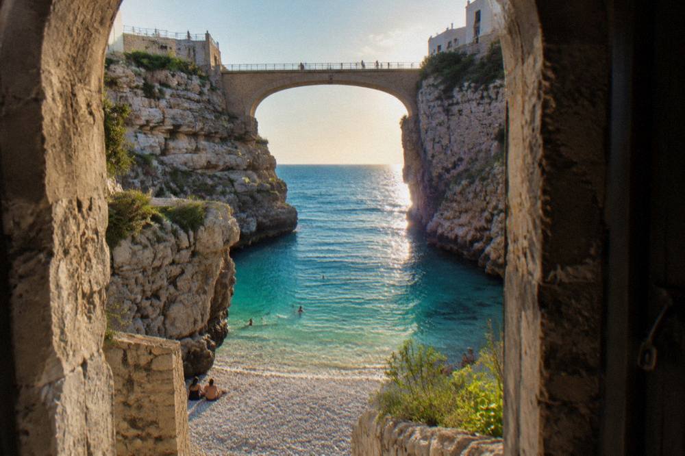 Narrow pebble cove between steep limestone cliffs under a stone arch bridge with turquoise water