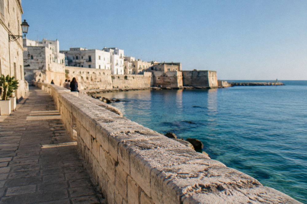 Sea promenade along an old town wall above calm turquoise water with distant people sitting on the wall