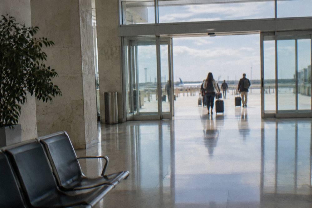 Arrivals hall with sliding glass doors on a bright day and distant travellers with wheeled suitcases