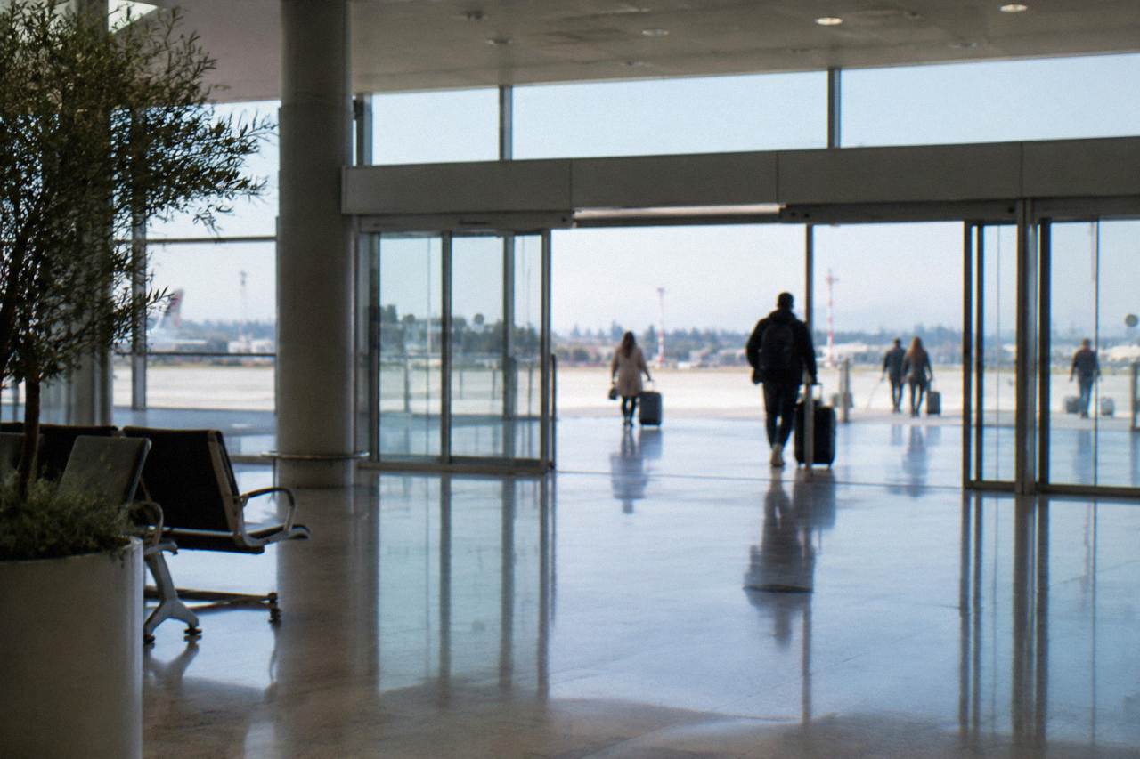 Arrivals hall with sliding glass doors on a bright day and distant travellers with wheeled suitcases