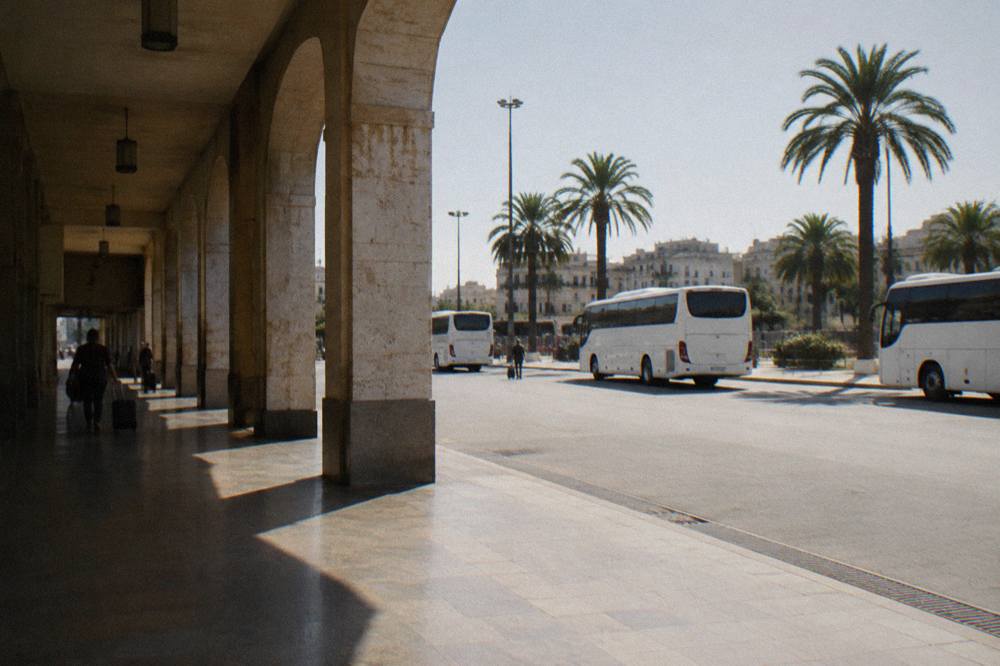 Station forecourt with plain white buses, palm trees and distant travellers with wheeled suitcases