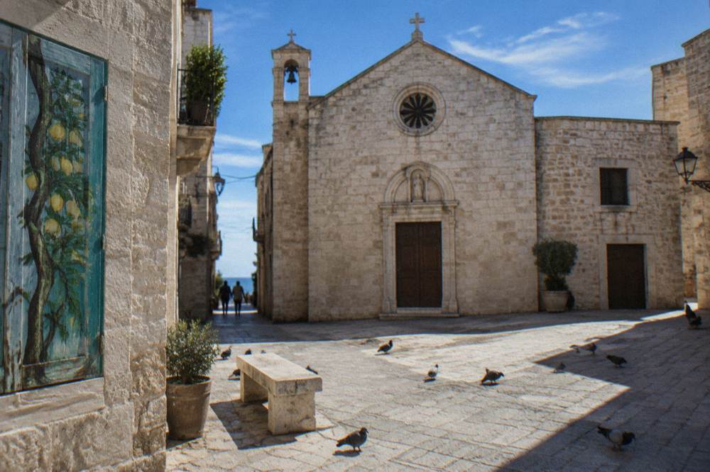 Small stone piazza with a modest pale limestone church front, a stone bench and pigeons in strong midday light