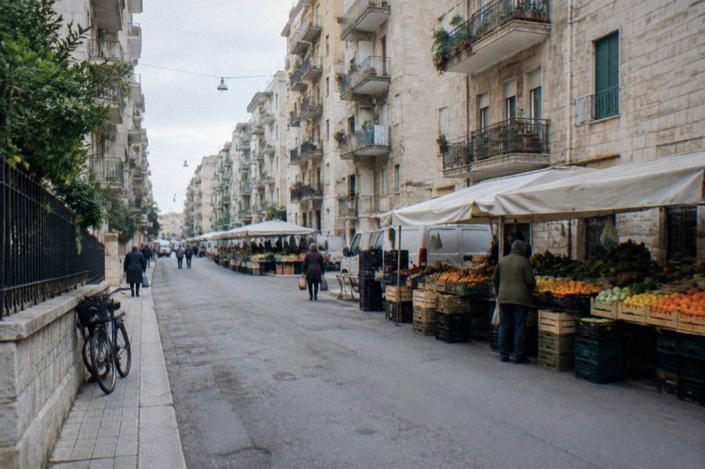 Residential street of apartment blocks with fruit and vegetable stalls under plain awnings and distant shoppers