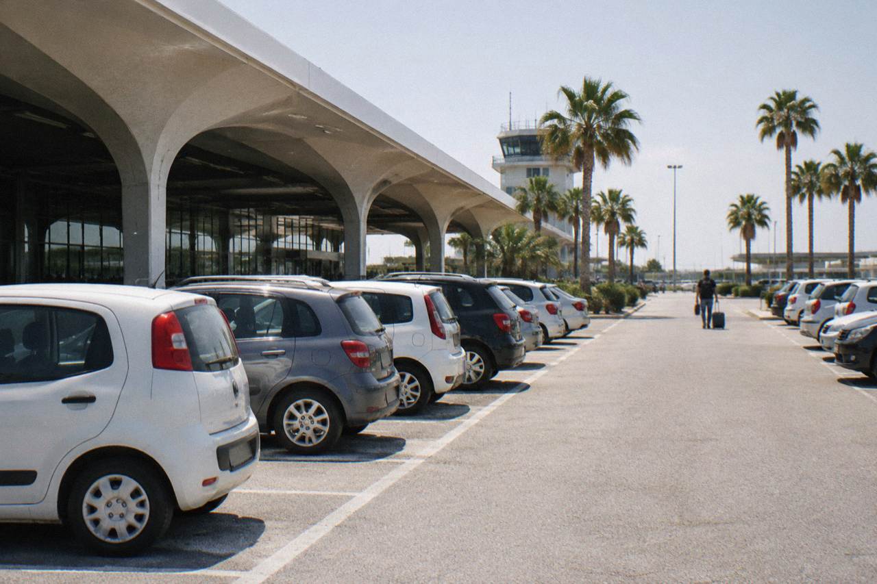 Rows of plain small rental cars in an airport car park with palm trees and a distant traveller with a suitcase