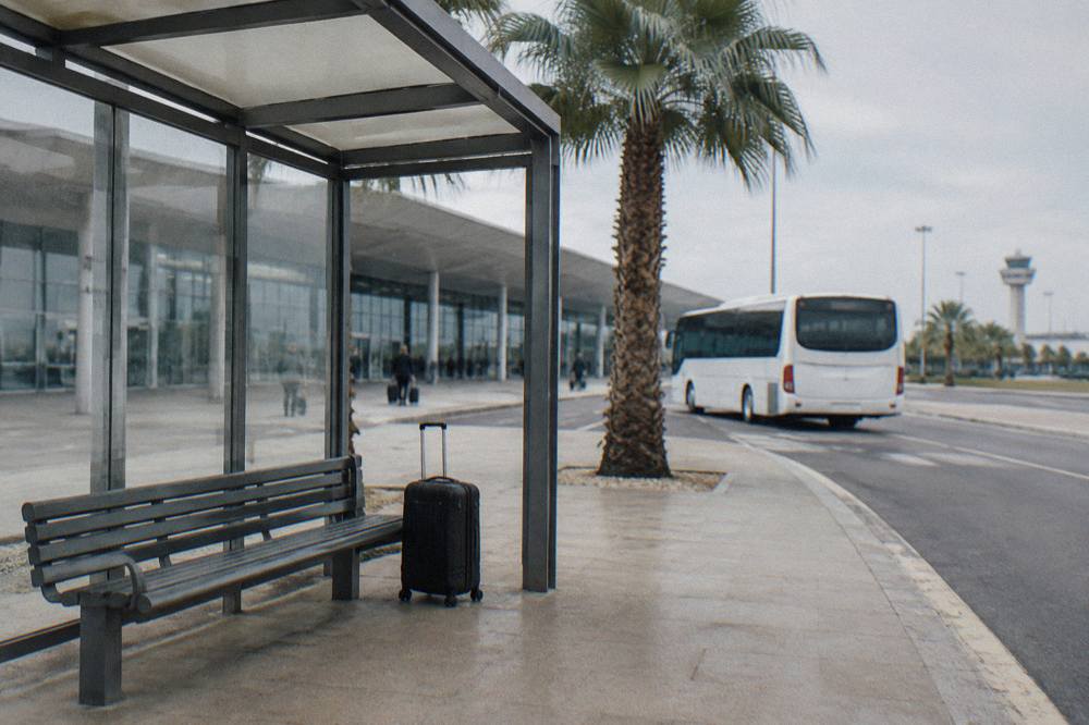 Bus shelter outside an airport terminal with a bench, a suitcase and a plain white bus waiting behind