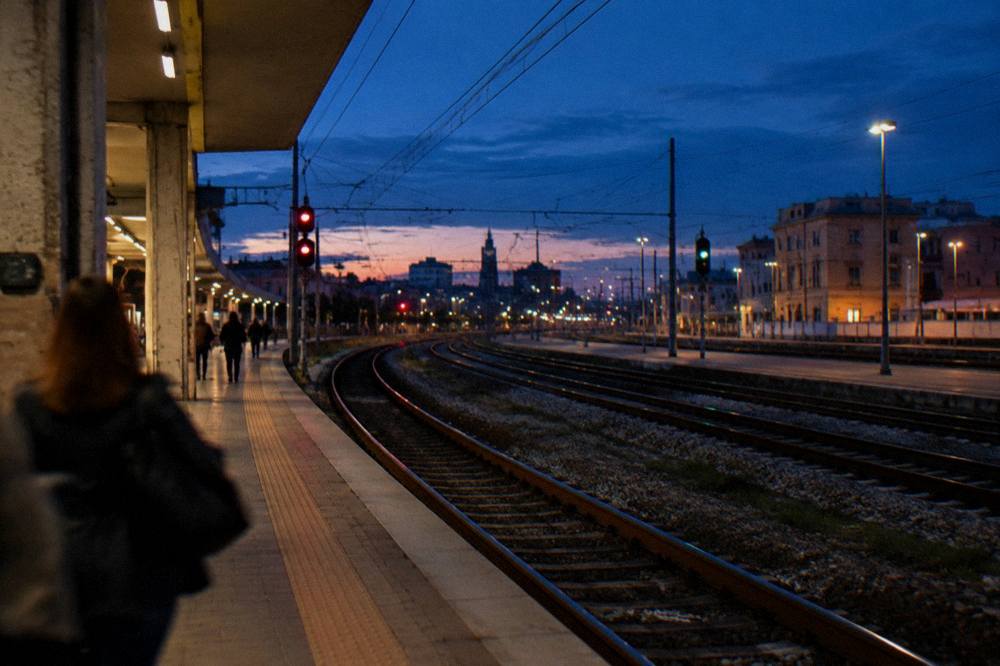 Rails curving away from a platform end at dusk with glowing signal lights and a few distant passengers