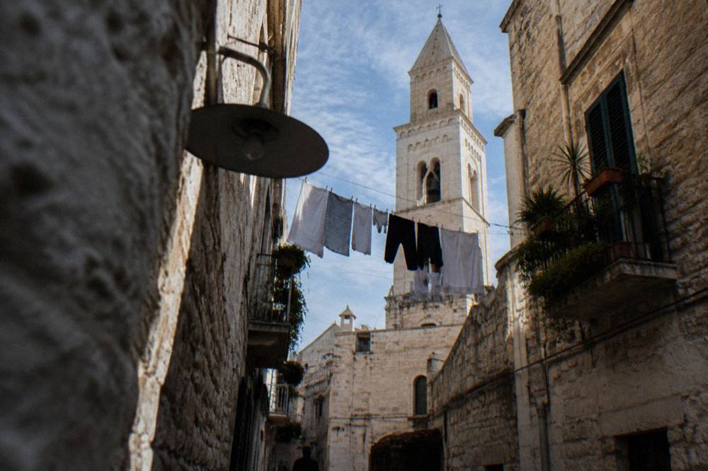 Looking up between close walls at a strip of blue sky crossed by washing lines and a bell tower