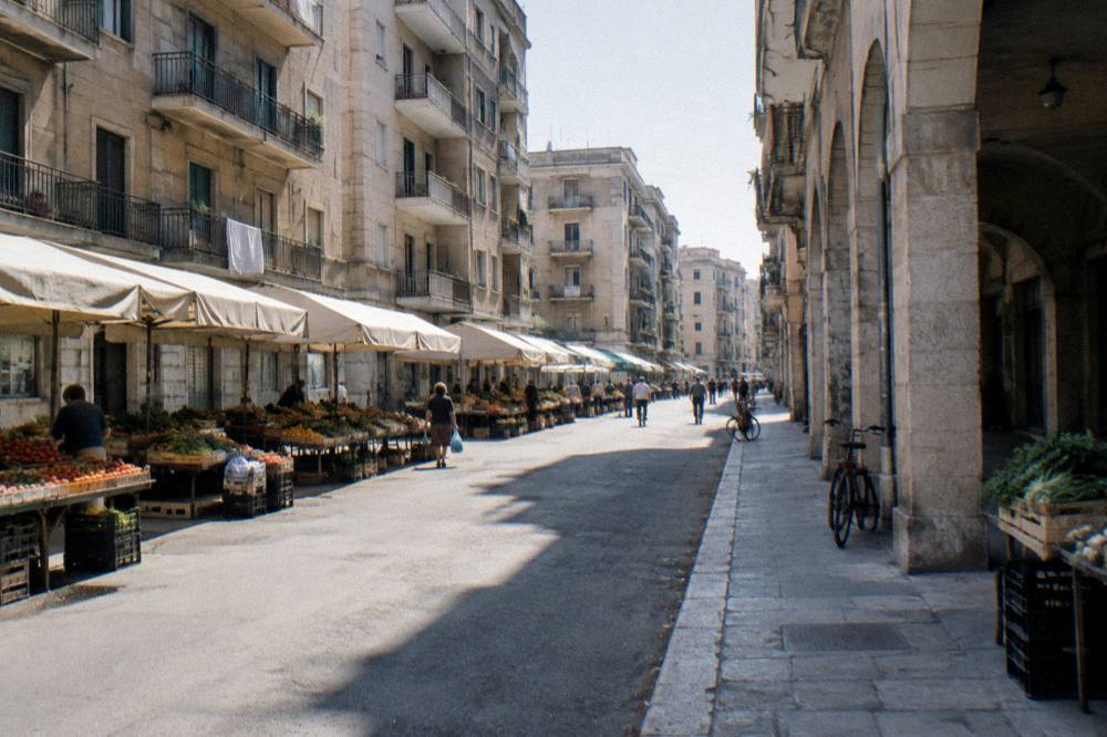 Residential street of apartment blocks with fruit and vegetable stalls under plain awnings and distant shoppers