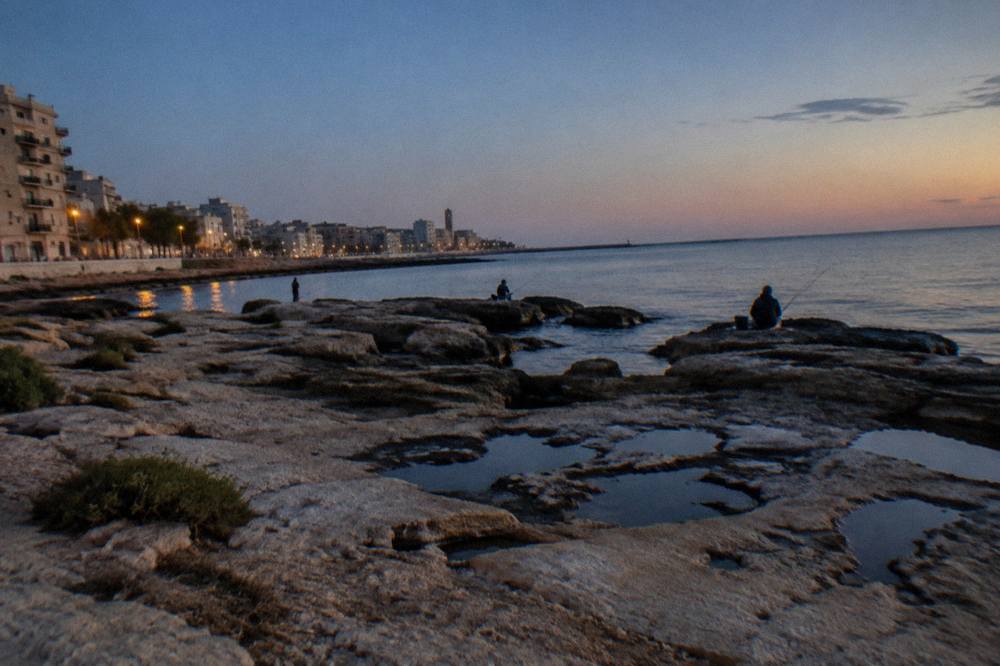 Seafront limestone rocks below a residential district with distant anglers and calm water in early evening
