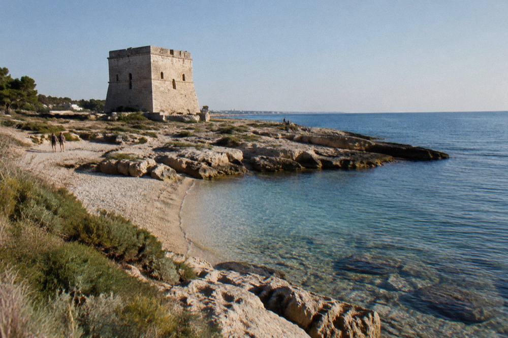Squat square stone watchtower on a flat rocky shore beside a small pebble beach and low scrub