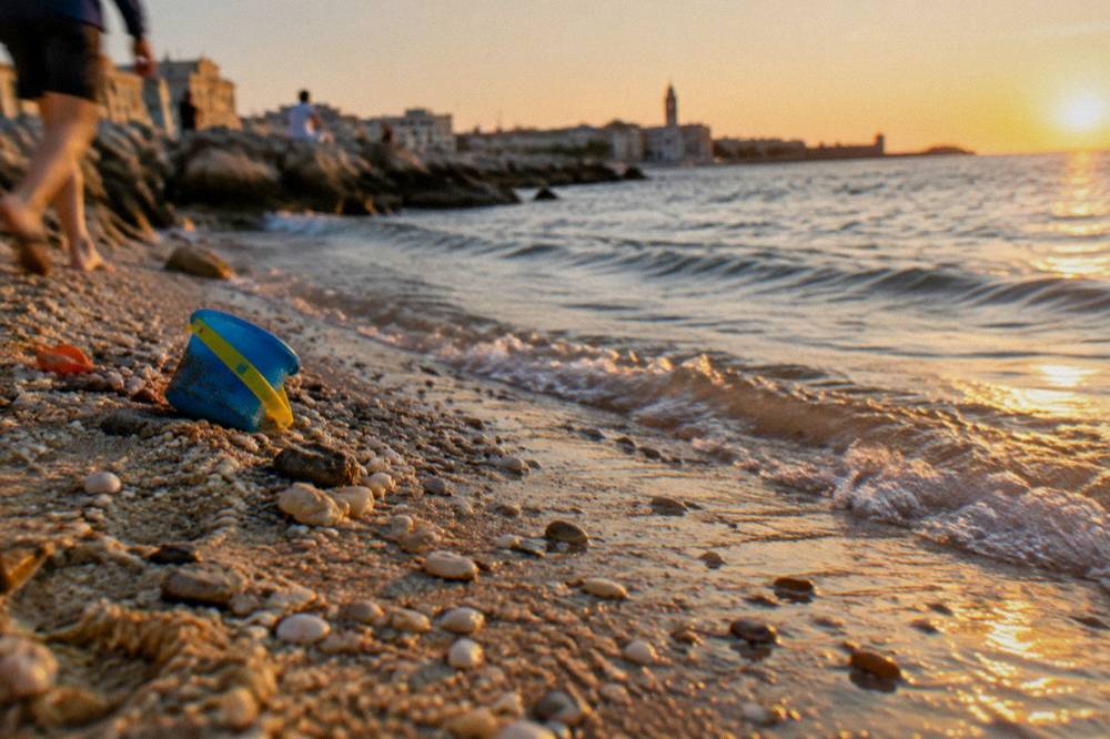 Pebbles and coarse sand at the water's edge with small clear waves and a plastic bucket left on the sand