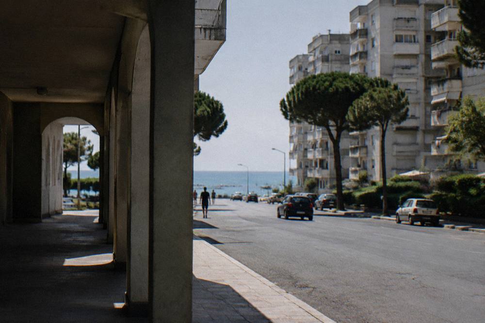 Residential district of 1960s apartment blocks with a wide road leading to a strip of sea between pine trees