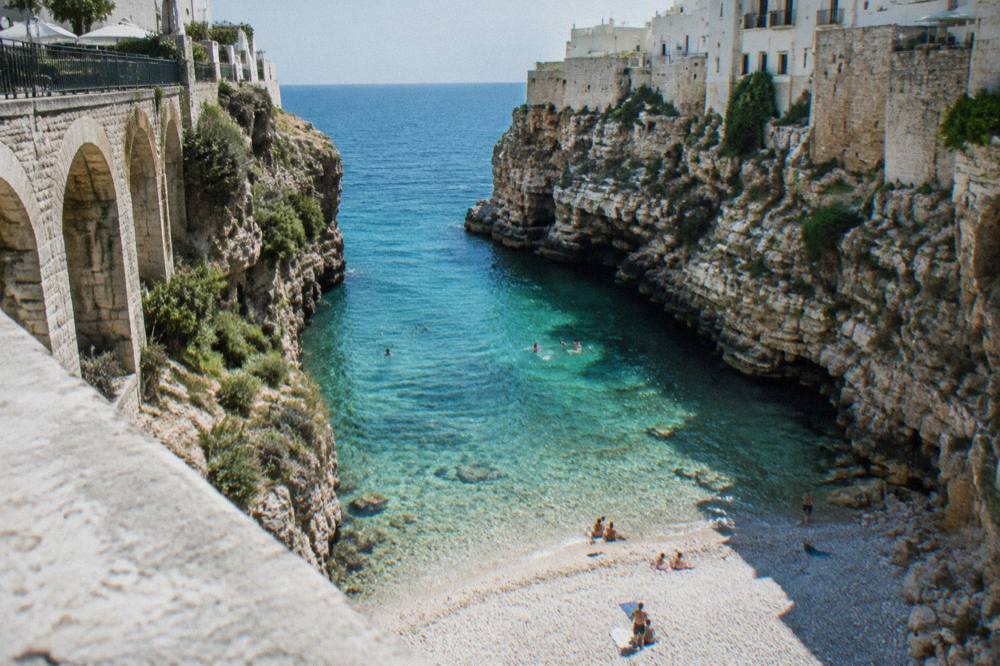View down from a stone bridge into a narrow cove of clear turquoise water and white pebbles