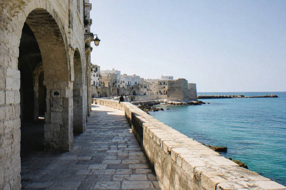 Sea promenade along an old town wall above calm turquoise water with distant people sitting on the wall