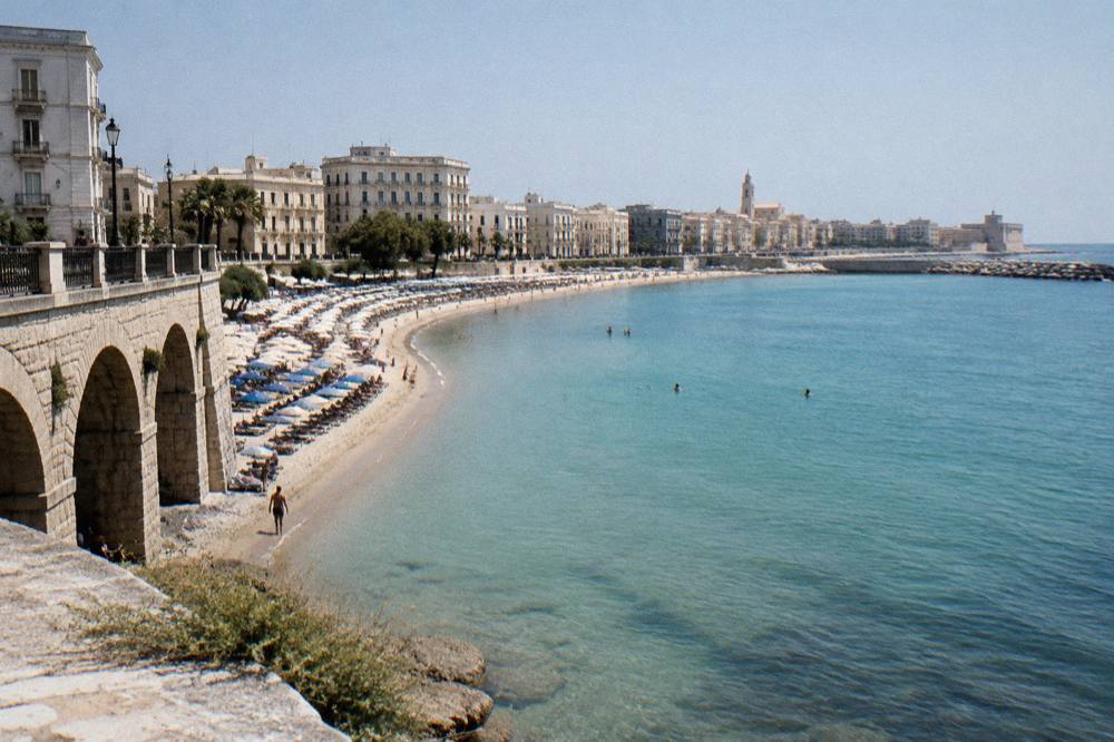 Small urban sandy beach with parasols in a curved bay, shallow turquoise water and seafront buildings behind