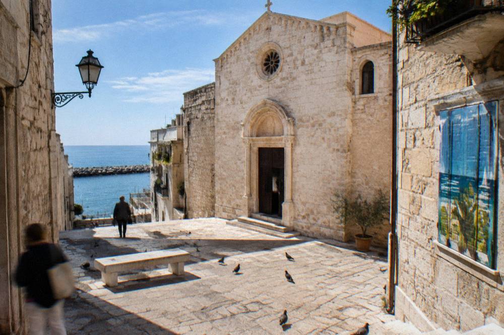 Small stone piazza with a modest pale limestone church front, a stone bench and pigeons in strong midday light