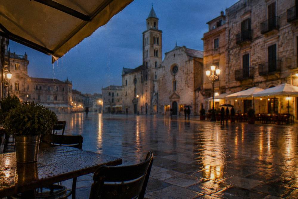 Rainy evening in a pale stone square with wet paving reflecting warm cafe lights and distant umbrellas