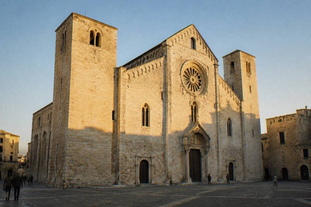 Romanesque church of pale limestone with a wide plain facade, two squat towers and a rose window above a paved square
