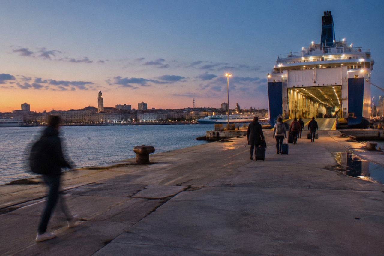 Distant passengers with wheeled suitcases walking along a quay toward a ferry's open loading ramp in evening light