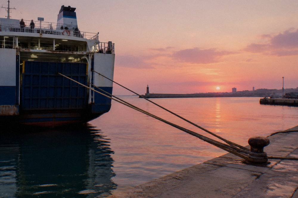 Stern of a ferry at a quay at sunrise with a pink sky, taut mooring ropes and still water