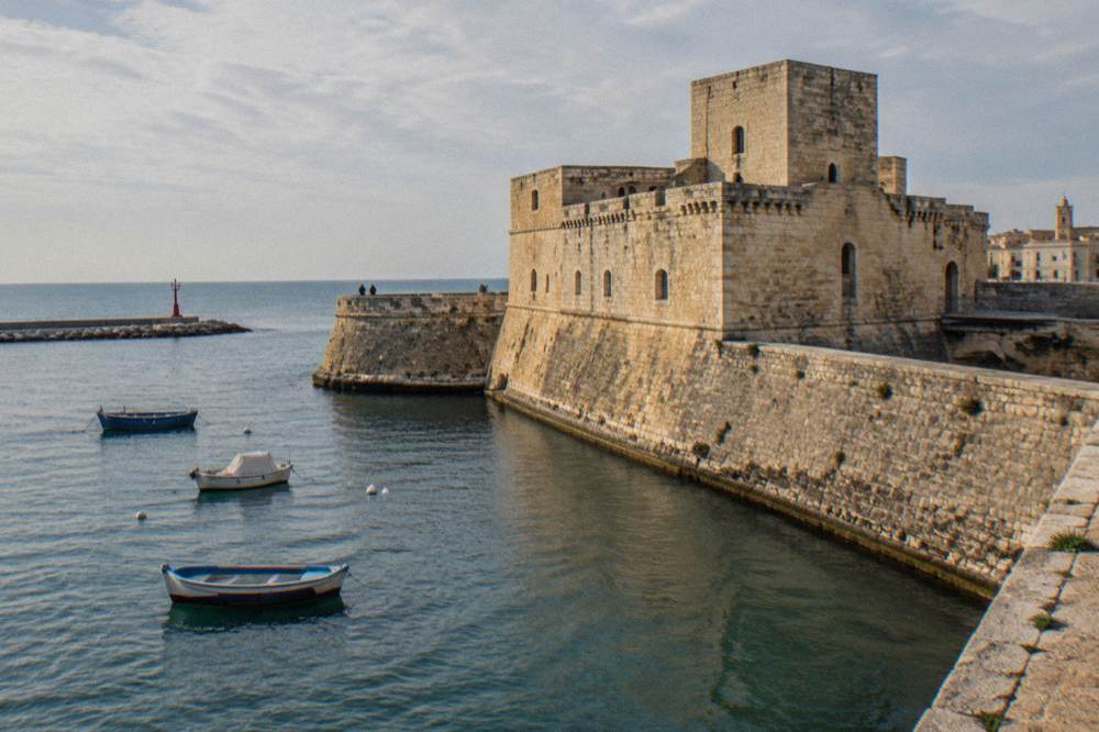Seaward wall of a pale stone castle beside harbour water with a few small boats in afternoon sun