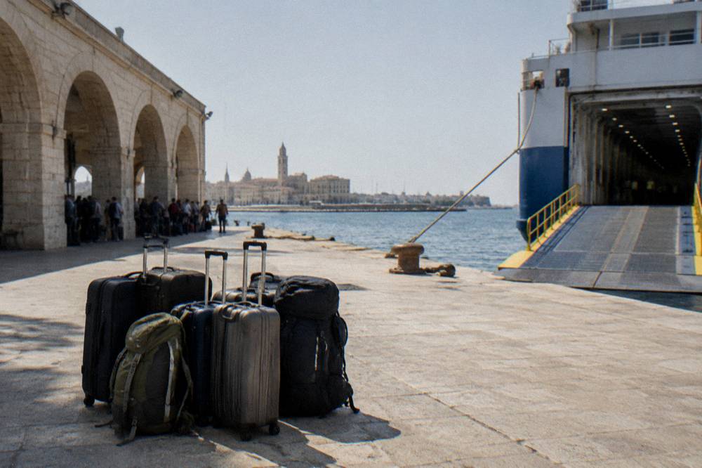 Suitcases and rucksacks stacked on a sunlit quay beside a ferry gangway with travellers waiting in the shade