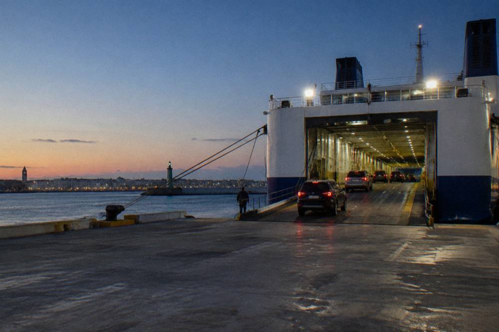 Ferry vehicle deck seen from the quay with rows of cars driving in under floodlights at evening