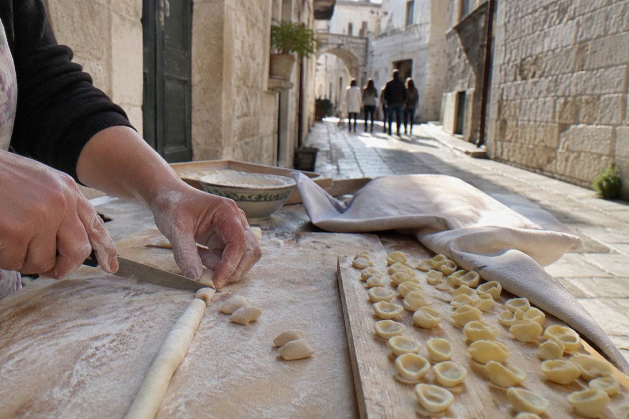 Floury hands drawing a small piece of dough across a board with a knife beside rows of finished pasta
