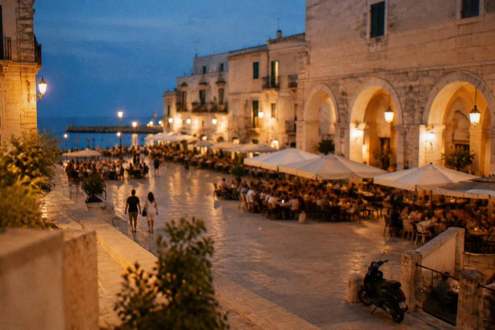 Evening piazza with warm light on cream walls, out-of-focus diners at tables and glowing market arches
