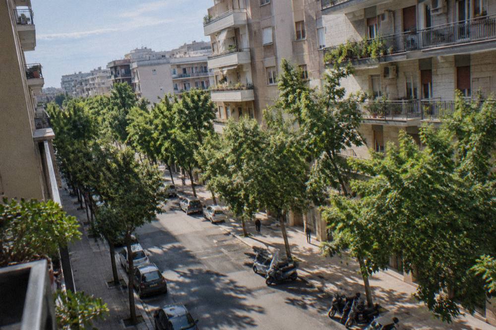 Leafy residential avenue of mid-century apartment blocks with street trees and parked scooters