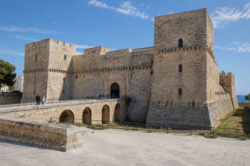 Massive square pale limestone fortress with corner towers, a dry moat and a bridge leading to an arched gate