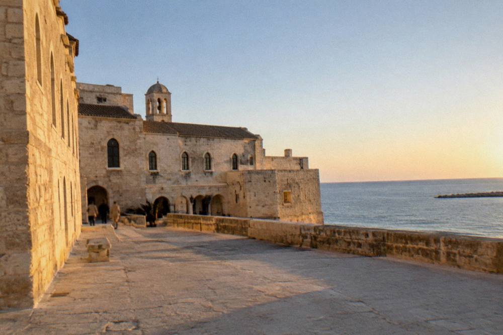 Low pale stone former monastery beside the sea with arched windows and a bare stone terrace