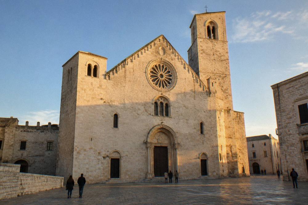Romanesque church of pale limestone with a wide plain facade, two squat towers and a rose window above a paved square