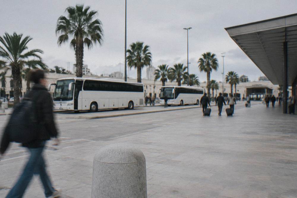 Station forecourt with plain white buses, palm trees and distant travellers with wheeled suitcases