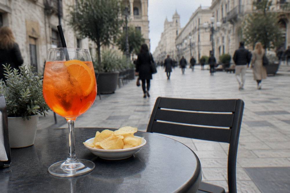 Pavement cafe table on a pedestrian street with an orange spritz and a small plate of crisps, passers-by blurred