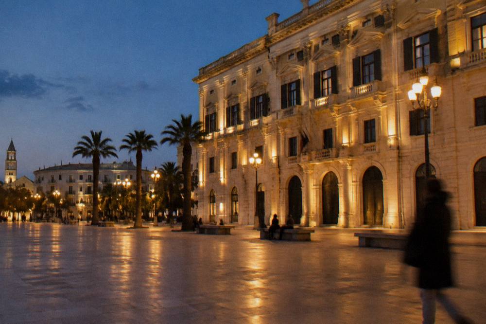 City square at evening with lit street lamps, distant people on benches and a warmly lit stone facade