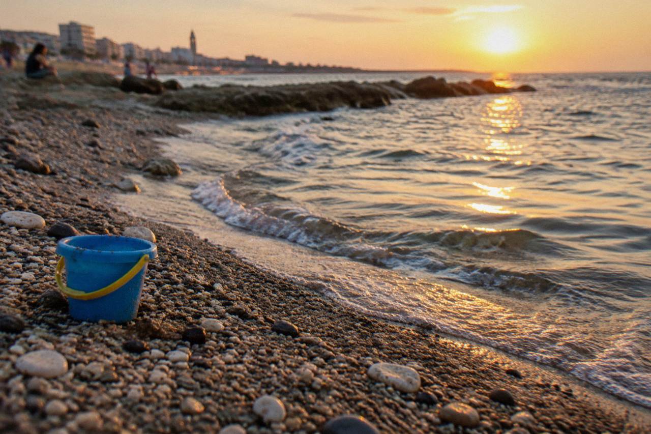 Pebbles and coarse sand at the water's edge with small clear waves and a plastic bucket left on the sand