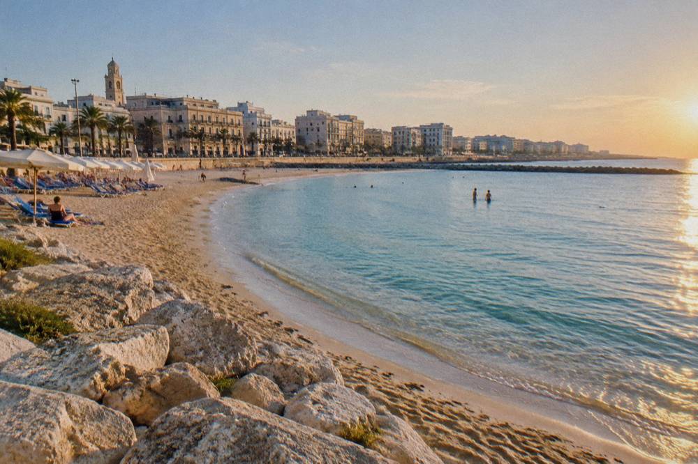 Small urban sandy beach with parasols in a curved bay, shallow turquoise water and seafront buildings behind