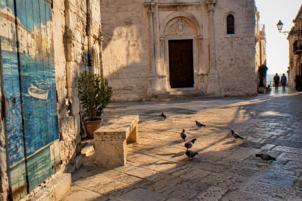 Small stone piazza with a modest pale limestone church front, a stone bench and pigeons in strong midday light