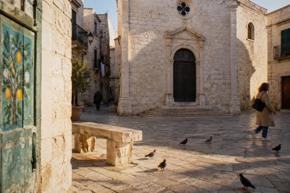 Small stone piazza with a modest pale limestone church front, a stone bench and pigeons in strong midday light