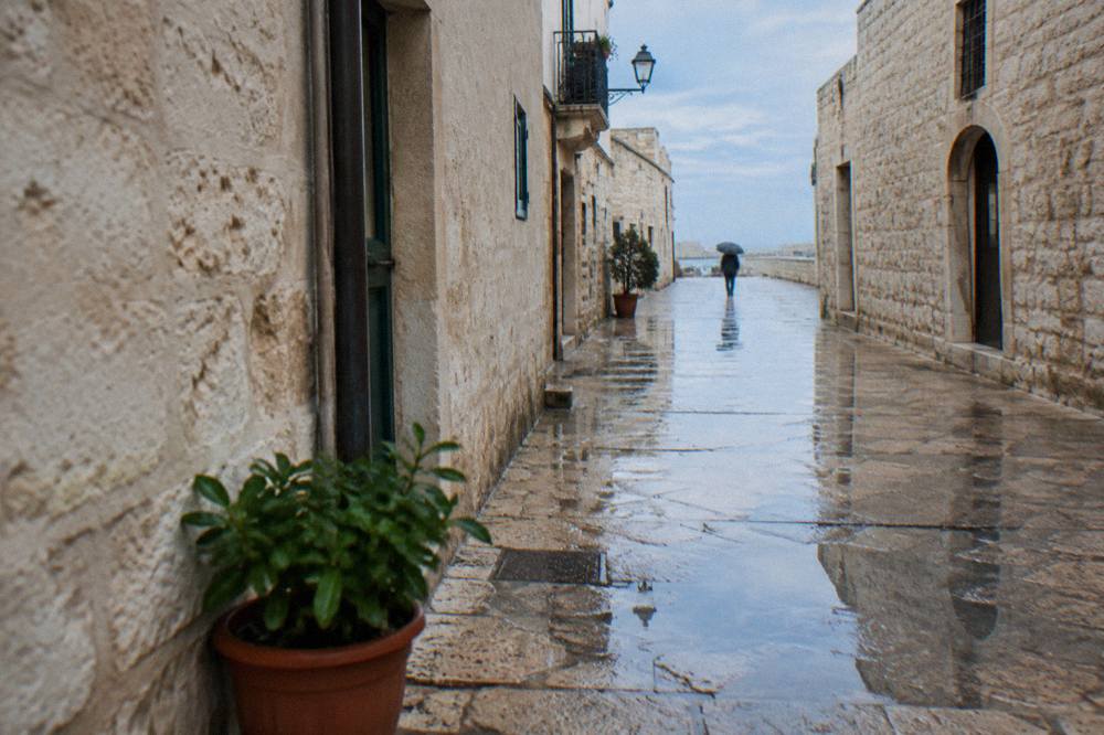 Wet limestone old town lane after rain with reflections in puddles and a distant person under an umbrella