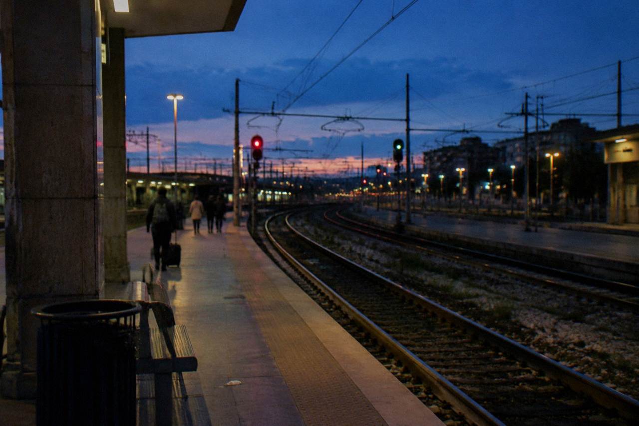 Rails curving away from a platform end at dusk with glowing signal lights and a few distant passengers