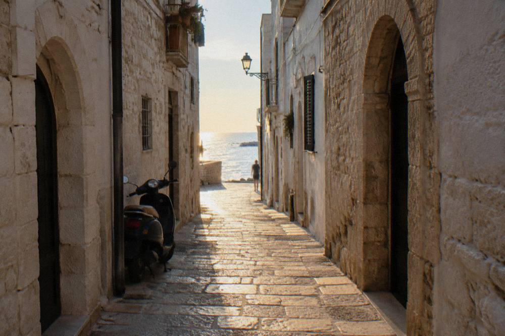 Straight old town lane between whitewashed walls ending in a bright slit of open sea with a parked scooter