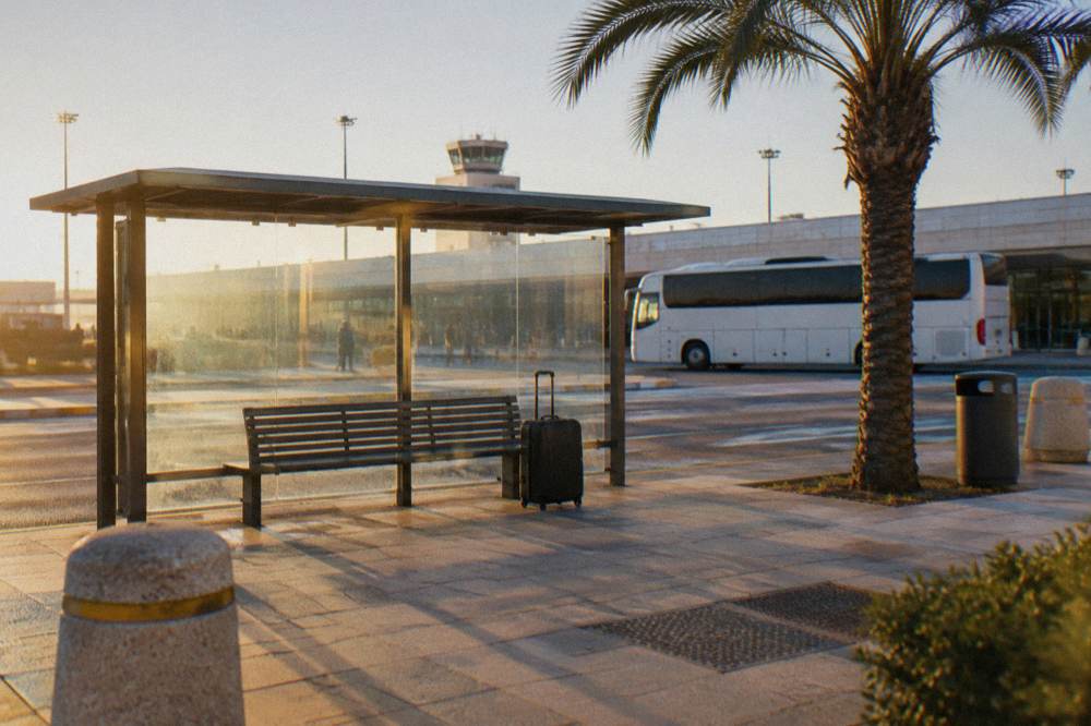Bus shelter outside an airport terminal with a bench, a suitcase and a plain white bus waiting behind